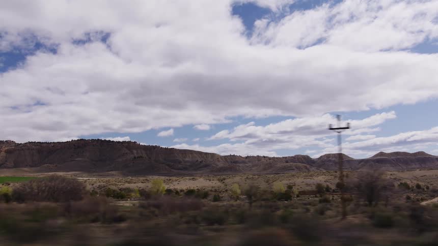 A passenger's point-of-view shot while driving past a vast, arid landscape with rolling hills and plateaus under a partly cloudy sky