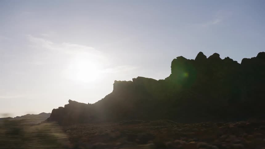 A cinematic side view from a moving car shows the sun setting behind rugged desert mountains