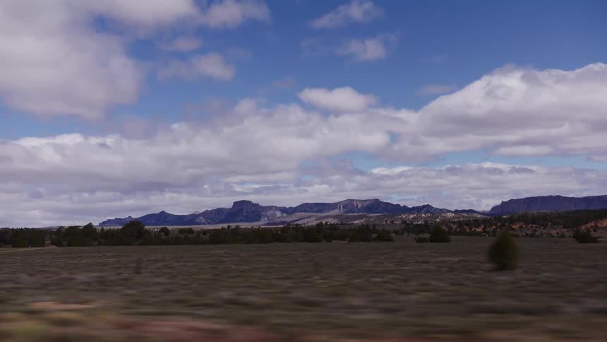 A slow-motion tracking shot reveals the vast, open high desert landscape with distant mountains under a beautiful cloudy blue sky