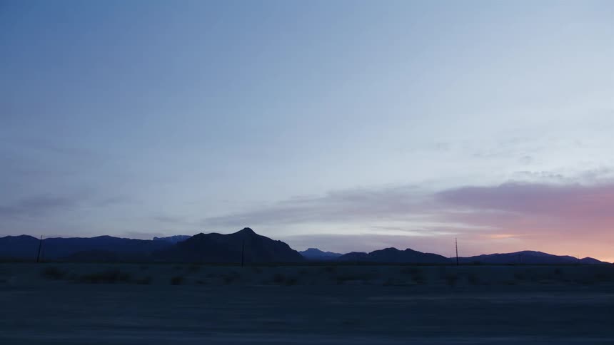 A serene view of a silhouetted desert mountain range against a beautiful sunrise sky from a moving vehicle