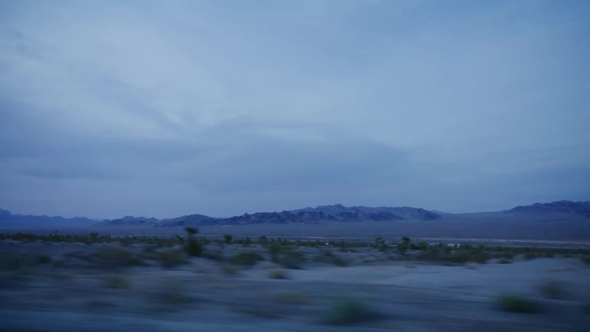 A serene drive through the vast, cool desert landscape at twilight with distant mountains under a cloudy sky