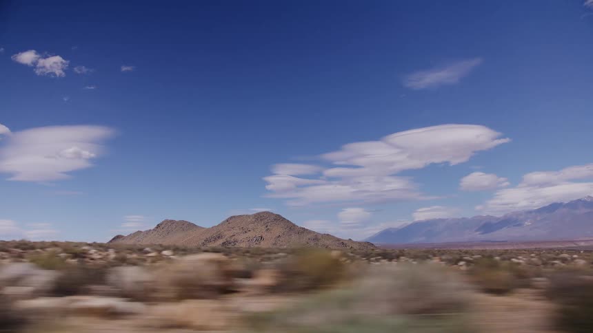 A scenic driving shot passes a vast desert landscape with rocky hills and distant mountains under a beautiful blue sky with scattered clouds