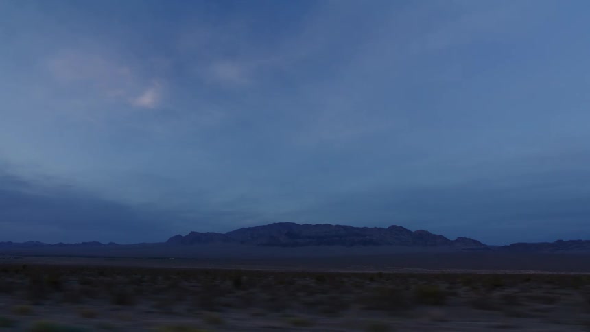 A serene driving shot passes a vast desert landscape with distant mountains under a tranquil blue twilight sky