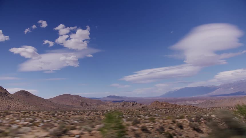 A view from a moving vehicle captures the vast, arid desert landscape and distant mountains under a bright blue sky with wispy clouds
