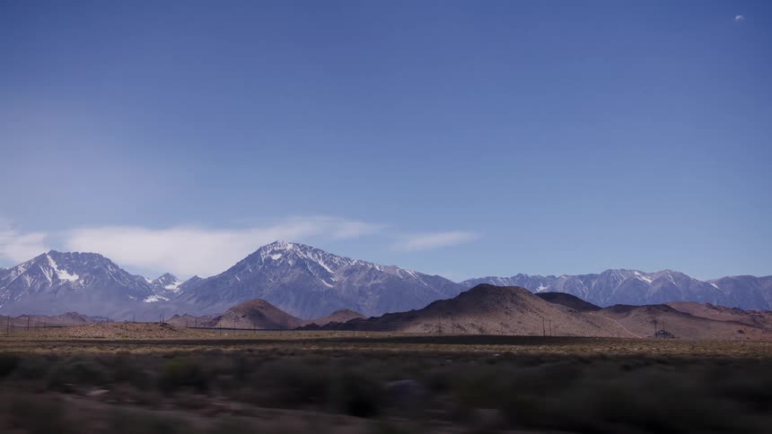 A scenic drive through a vast desert valley with a majestic snow-capped mountain range in the distance under a clear blue sky