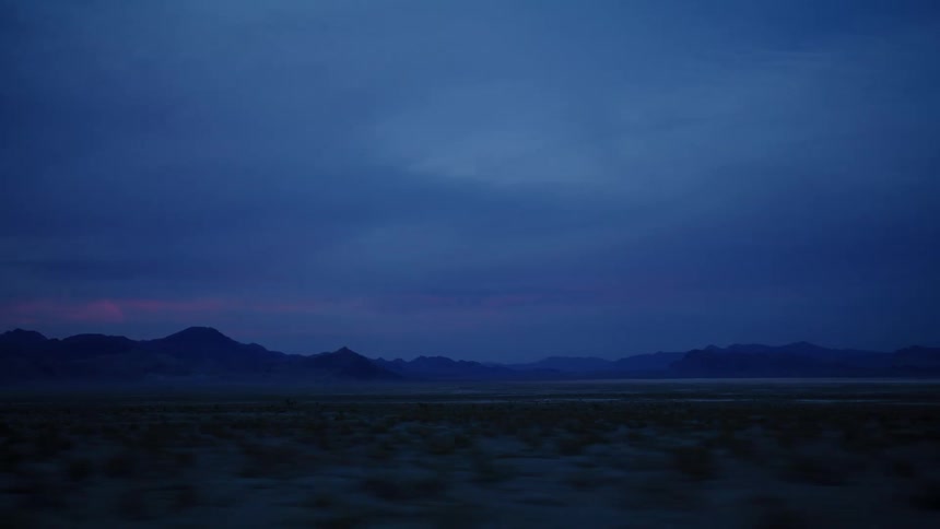 Driving through a vast desert landscape at twilight as mountains are silhouetted against a dark blue and pink sky