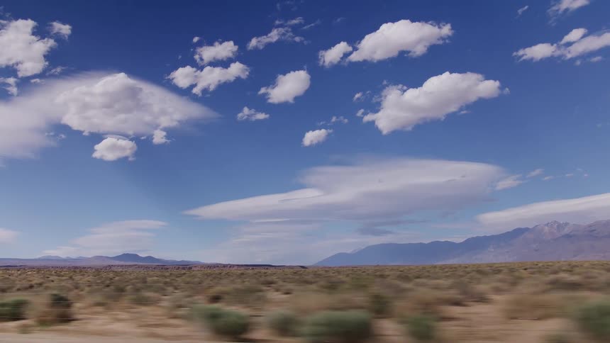 A scenic side-view driving plate through the vast desert landscape of Death Valley with mountains in the distance under a clear blue sky