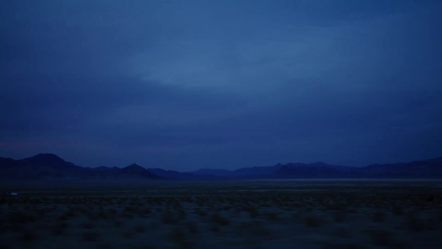 A moody view from a moving car captures the vast desert landscape and distant mountains during the blue hour at dusk
