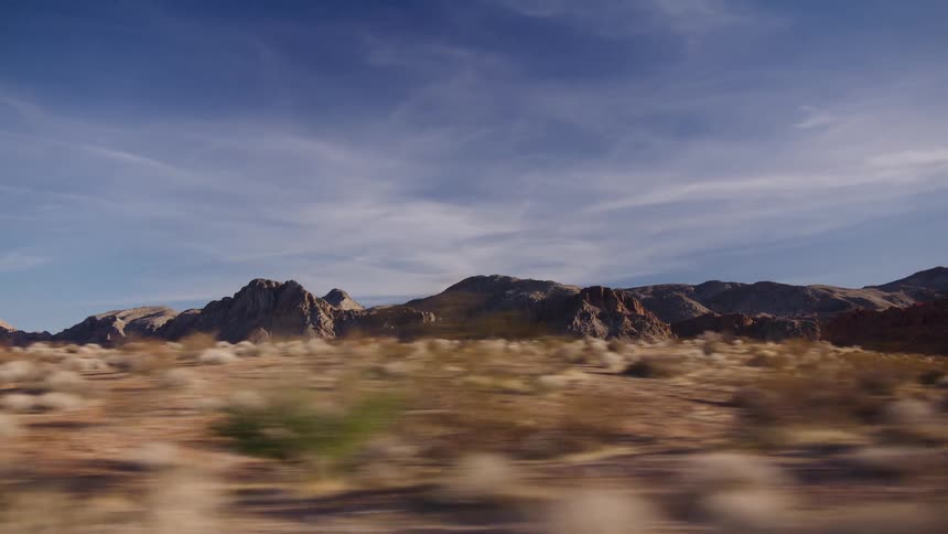 A beautiful desert landscape with rocky mountains and a clear blue sky speeds by from the point of view of a car window