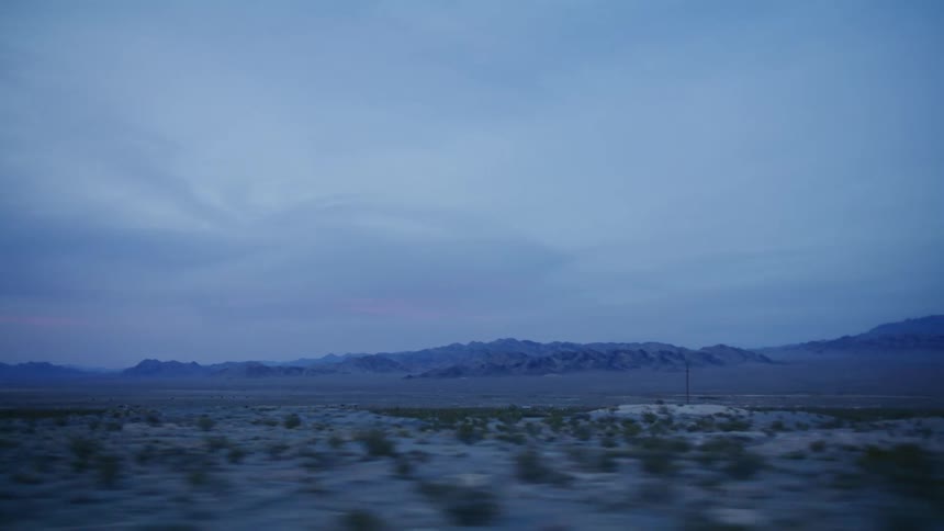 Driving through a vast and desolate desert landscape with distant mountains under a moody twilight sky