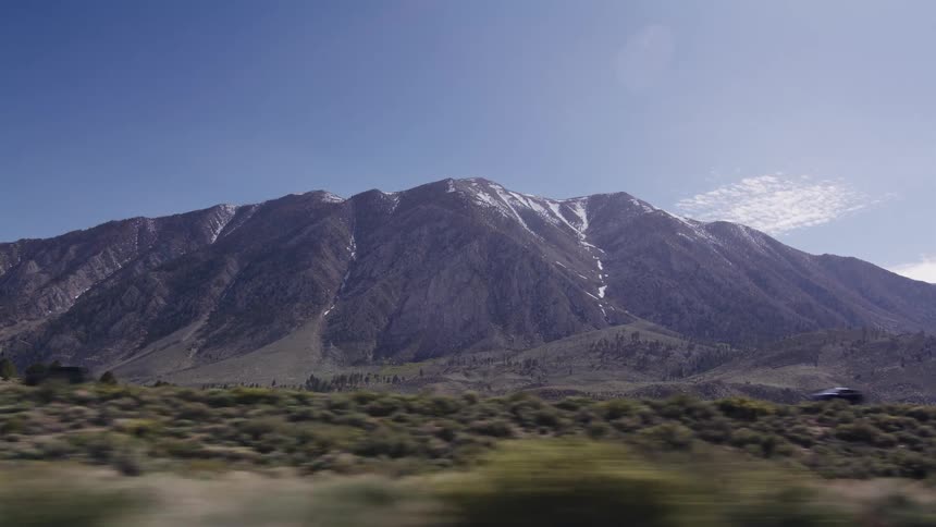 A slow-motion driving shot reveals a majestic mountain with snow-capped peaks under a clear blue sky