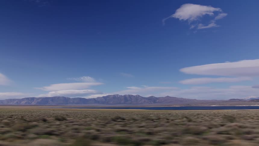 A scenic vehicle window view of a vast desert plain, a distant lake, and rugged mountains under a clear blue sky