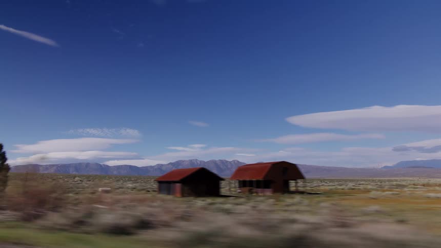 A scenic drive through a wide-open valley, passing old rustic barns with a mountain range on the horizon under a clear blue sky