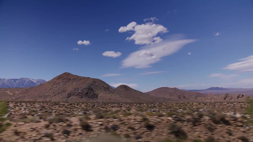 A side-view driving plate captures the arid desert landscape with rocky hills and snow-capped mountains in the distance under a clear blue sky