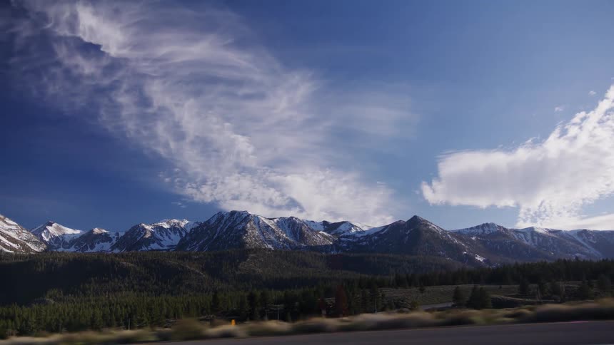A scenic drive reveals the majestic snow-capped Sierra Nevada mountains under a vast blue sky with wispy clouds
