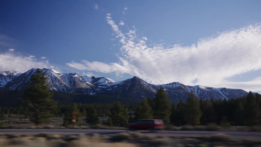 A scenic view from a moving car captures the majestic snow-capped mountains and pine tree forest under a beautiful blue sky
