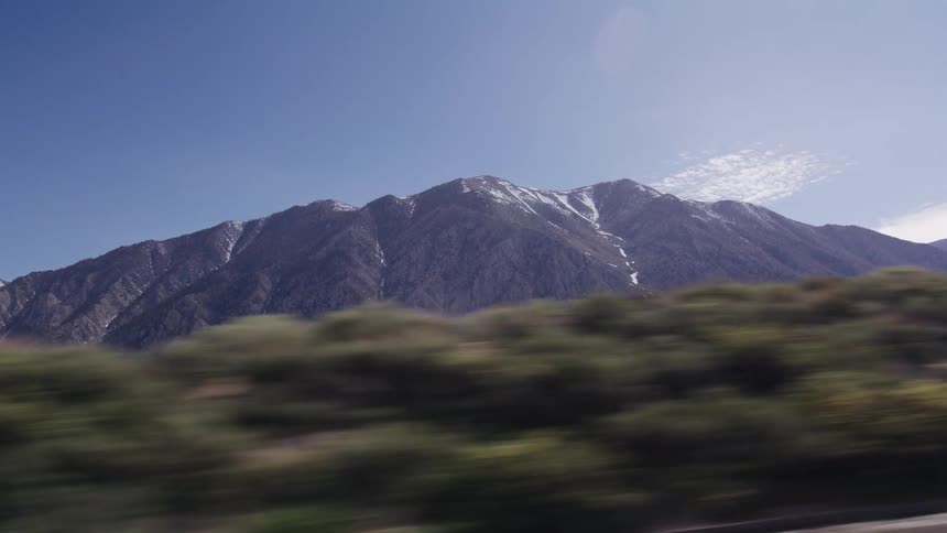 Passenger's point of view driving past a vast, snow-capped mountain range on a sunny day