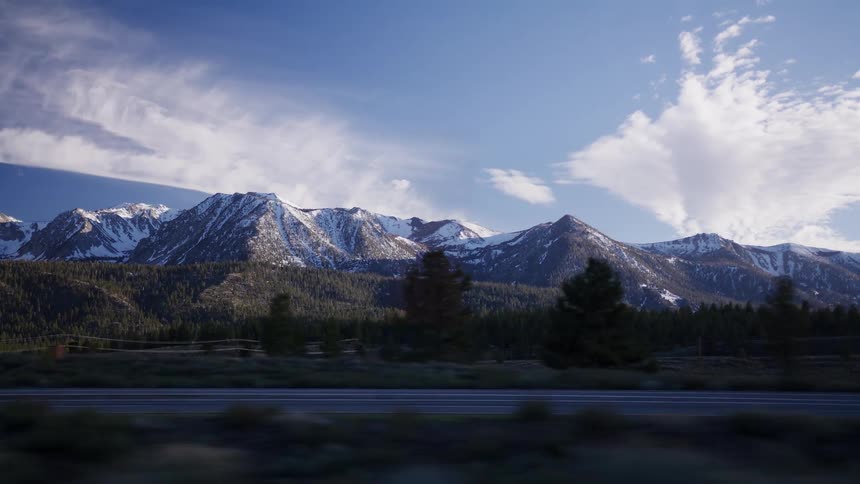 A scenic driving shot reveals a majestic, snow-capped mountain range behind a lush green forest