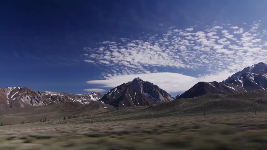 A scenic drive past a vast, snow-capped mountain range under a deep blue sky with wispy clouds
