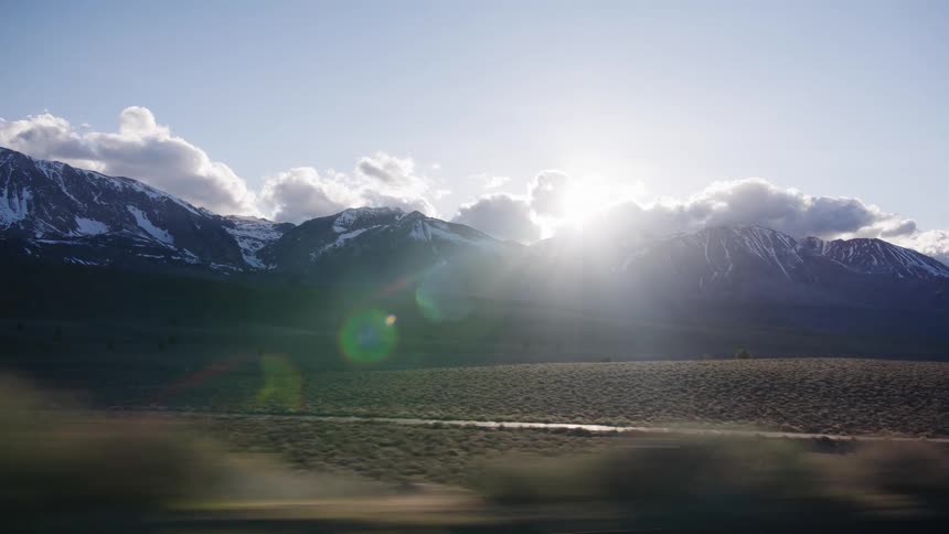 A beautiful scenic drive passes snow-capped mountains as the sun peeks out from behind the clouds creating a dramatic lens flare
