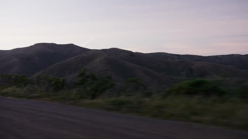 A serene view from a moving car shows rolling hills and a dirt road at dusk