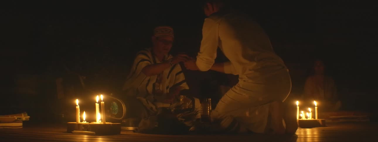 A shaman in traditional attire gives a sacred ceremonial drink to a man during a spiritual ritual by candlelight
