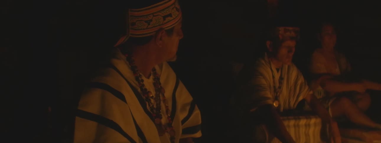 Indigenous men in traditional clothing and beaded necklaces sit together during a spiritual fire ceremony at night