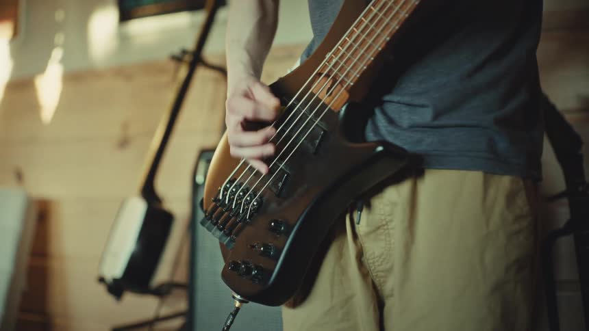 Close-up of a musician's hands as they energetically strum a five-string electric bass guitar with a pick in a sunlit music room