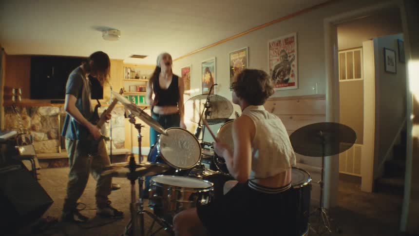 An energetic teenage band with a female singer, guitarist, and drummer rock out during a rehearsal in a basement