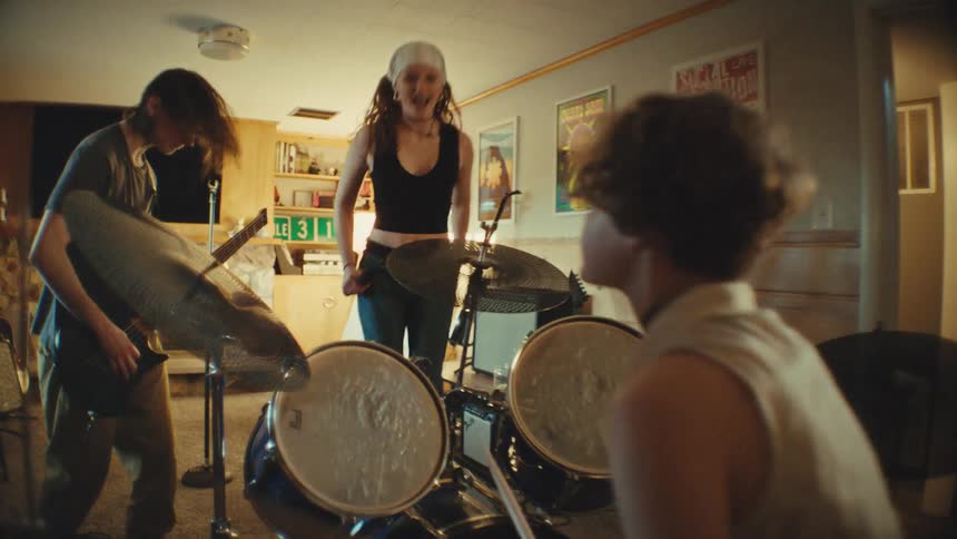 An energetic teenage band with a female singer, guitarist, and drummer rock out during a rehearsal in a basement