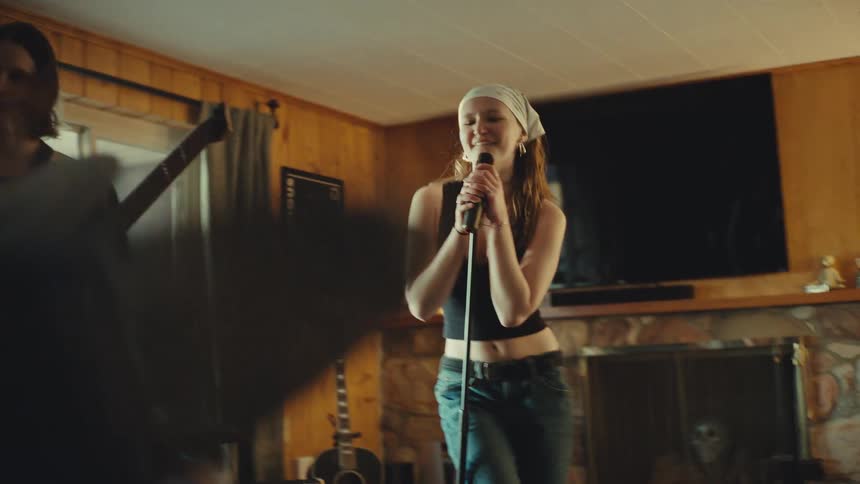 A young woman headbangs and sings into a microphone while her band practices in a rustic living room