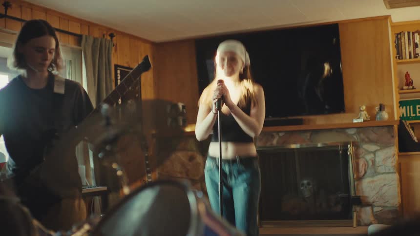 A young woman headbangs and sings into a microphone while her band practices in a rustic living room