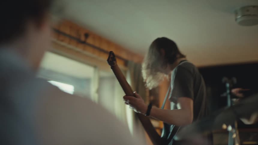 From behind a drum kit, a long-haired guitarist shreds on his electric guitar while a female singer performs during an energetic band practice