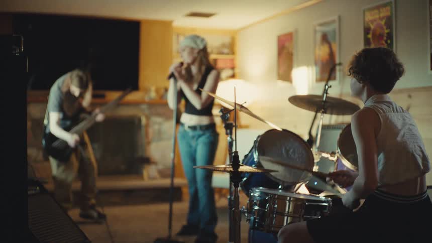 A young rock band with a female vocalist and drummer passionately rehearses with their male guitarist in a cozy basement
