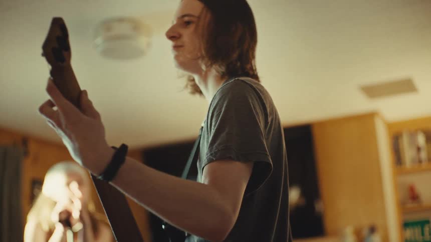 A long-haired teenage boy intensely plays an electric guitar while practicing with his band in a living room