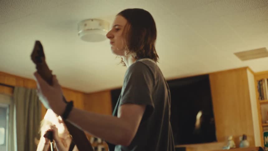 A long-haired teenage boy intensely plays an electric guitar while practicing with his band in a living room