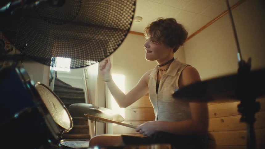 A passionate young drummer smiles while playing a groovy beat on their drum kit in a hazy, sun-drenched room