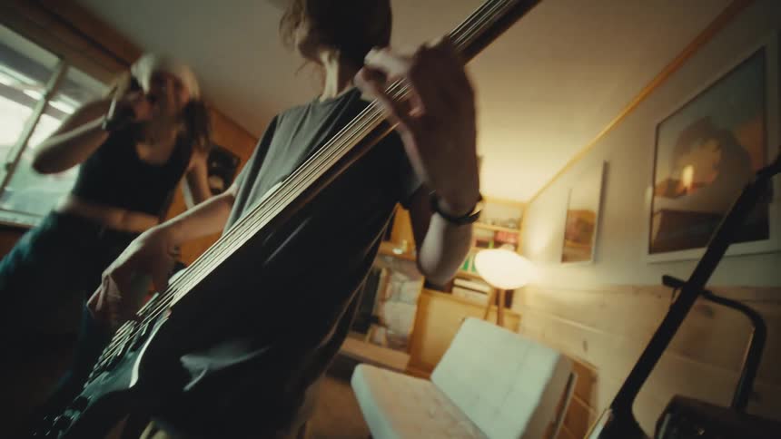 A dynamic low-angle view shows a bassist energetically playing a black bass guitar while a female singer performs in their cozy home music studio