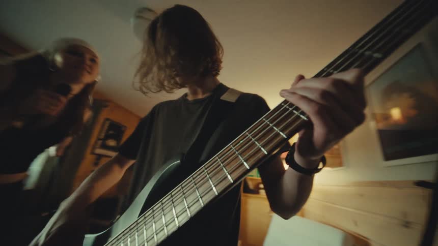 A dynamic low-angle view shows a bassist energetically playing a black bass guitar while a female singer performs in their cozy home music studio