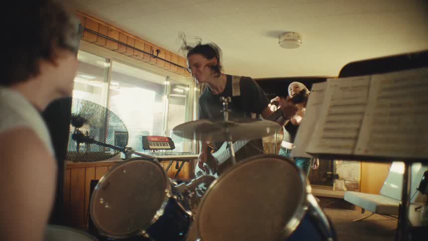 Cool young band with a female vocalist and guitarist rehearsing their music with energy, viewed from behind the drummer