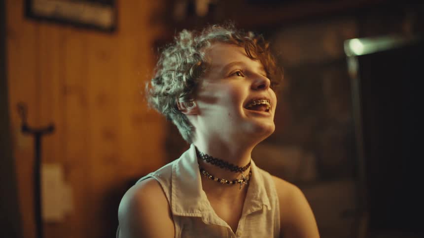 A happy young person with braces and curly hair looks up in pure joy and wonder inside a cozy room