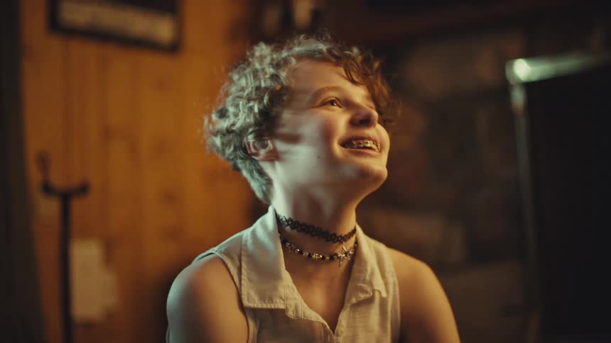 A happy young person with braces and curly hair looks up in pure joy and wonder inside a cozy room