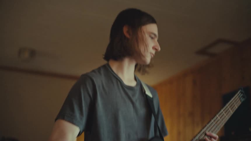 Focused young musician with medium-length hair plays a fast riff on his five-string electric bass guitar in a practice room