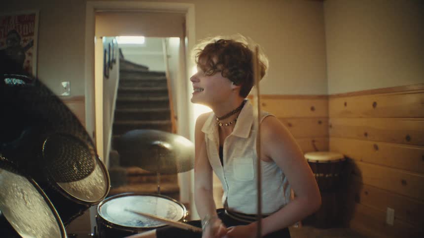 A joyful young drummer with short hair plays a beat on their electronic drum kit in a warm, sun-drenched music room