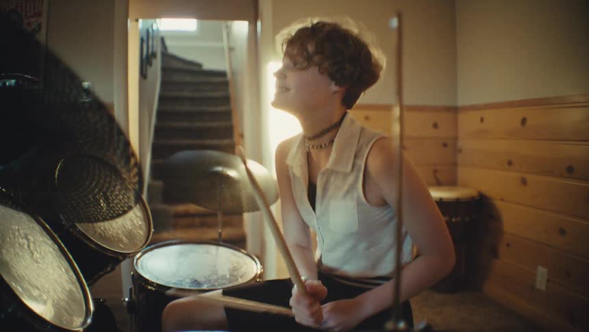 A joyful young drummer with short hair plays a beat on their electronic drum kit in a warm, sun-drenched music room