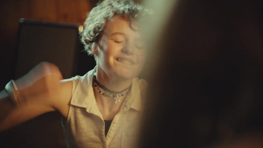 An energetic teenage girl with curly hair and braces laughs and dances with excitement while talking to a friend