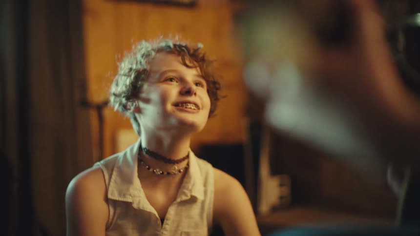 A cheerful teenager with curly hair and braces smiles brightly while engaged in a conversation in a cozy, warm room