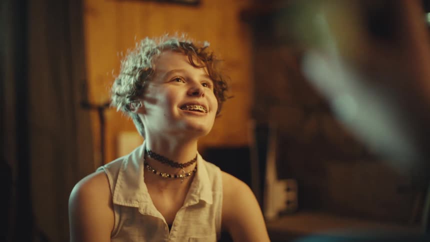 A cheerful teenager with curly hair and braces smiles brightly while engaged in a conversation in a cozy, warm room