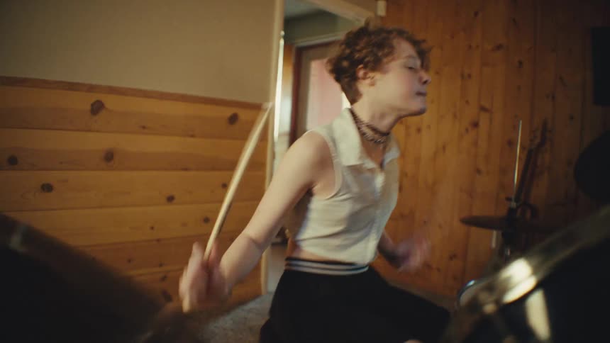 A young rock drummer passionately plays the drums in a wood-paneled home studio, with the shot racking focus to a close-up of the cymbal
