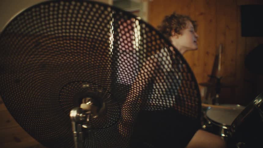 A young rock drummer passionately plays the drums in a wood-paneled home studio, with the shot racking focus to a close-up of the cymbal